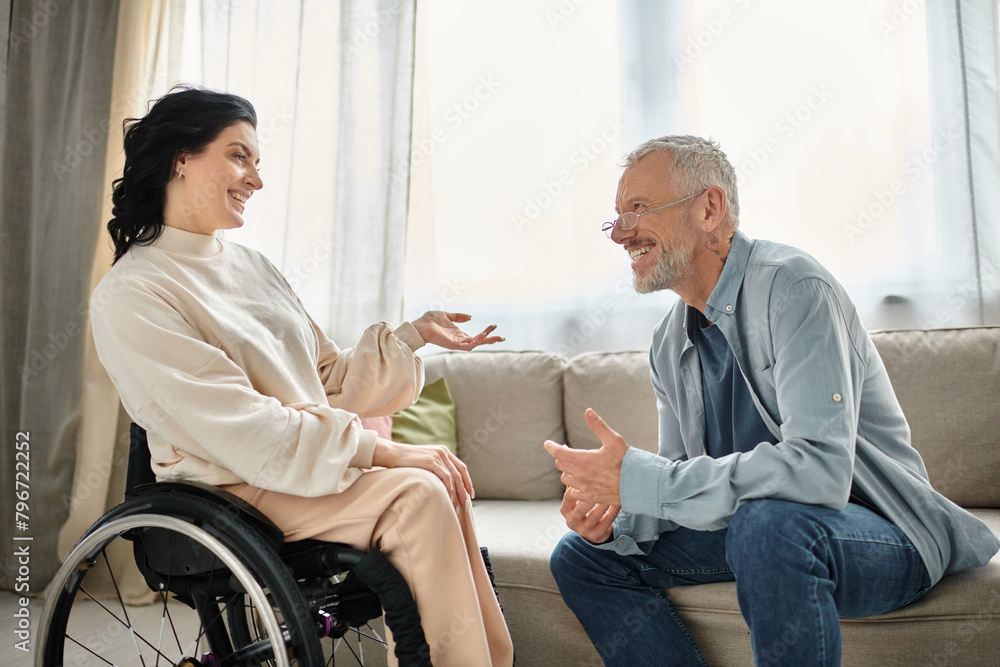 A man converses with a disabled woman in wheelchair in a cozy living room setting.