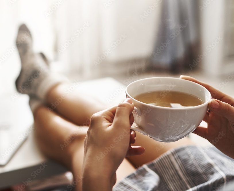 Cup of tea and chill. Woman lying on couch, holding legs on coffee table, drinking hot coffee and enjoying morning, being in dreamy and relaxed mood. Girl in oversized shirt takes break at home